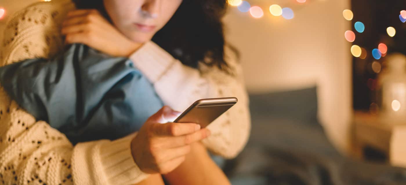 A woman sitting on a bed and using a cell phone. There are holiday lights in the background.