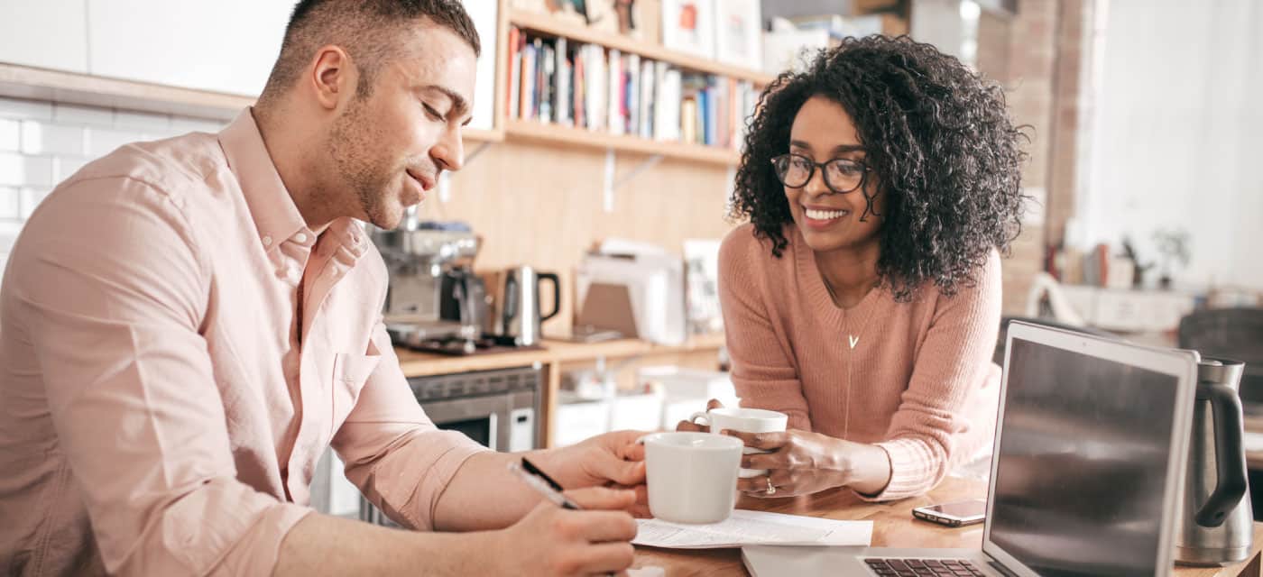 a couple drinking out of mugs, they are sitting at a table with a laptop.