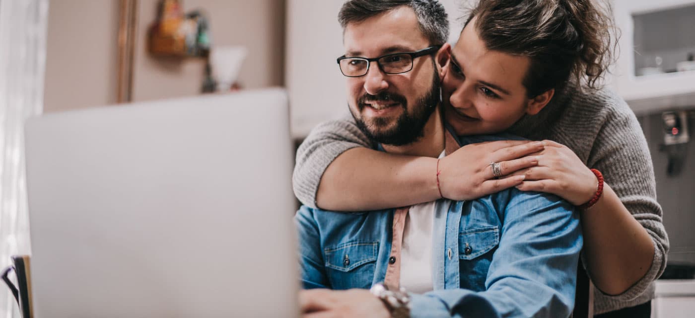 A couple using a laptop. The man is sitting at a table and the woman is hugging him from behind.