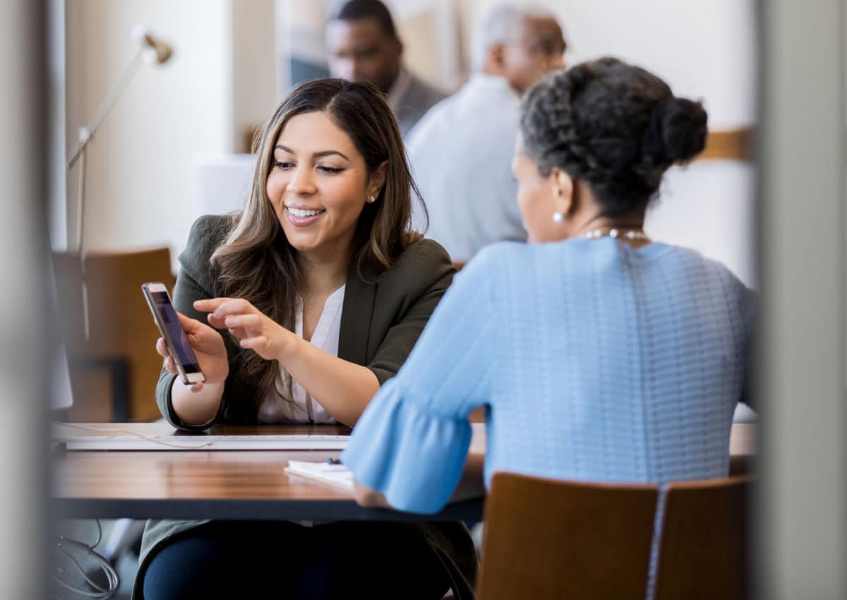 Two women sitting at a table wearing business clothing, one of the women is showing the other one something on a phone.