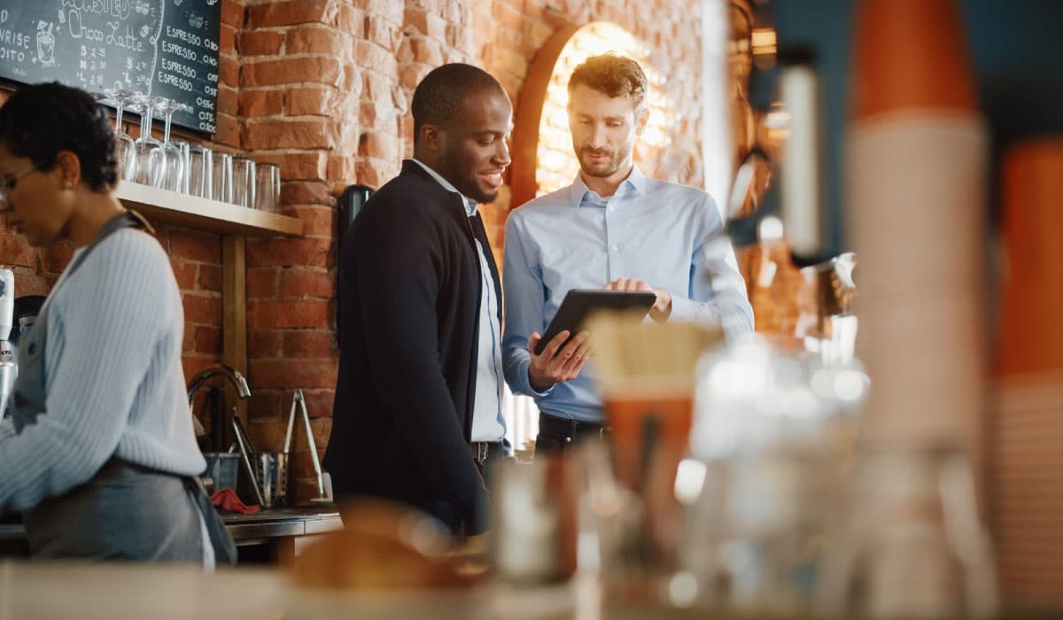 Two men standing and looking at a tablet. They are at a bar.