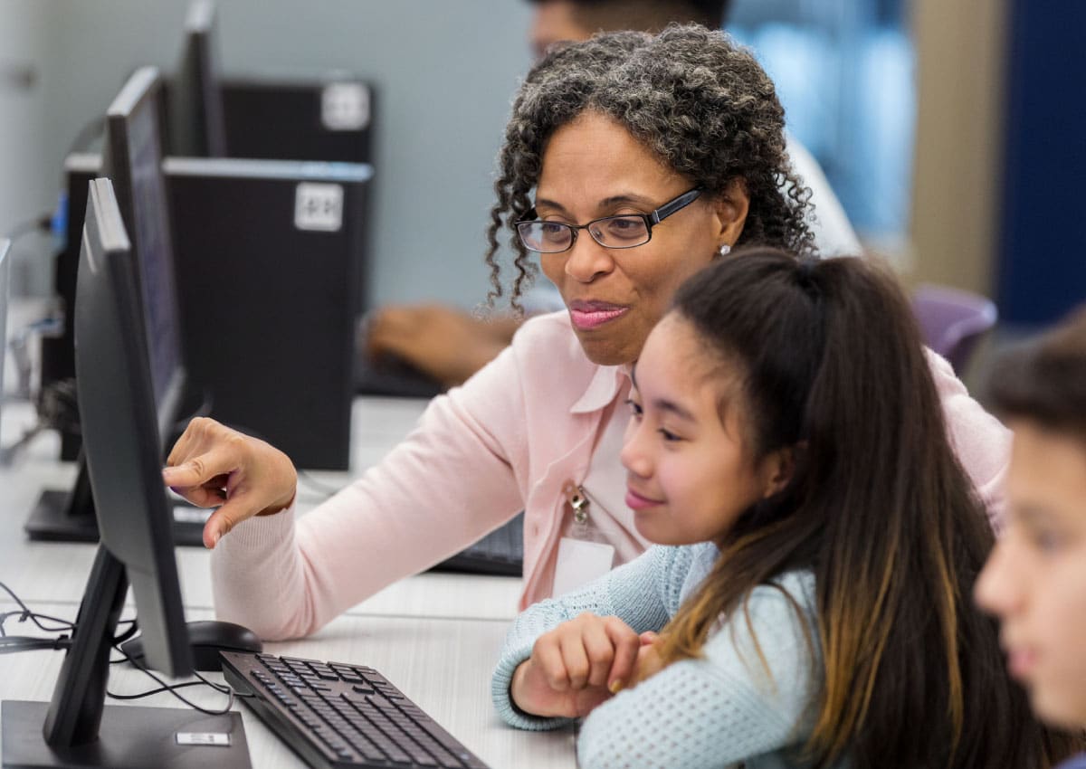 A young girl looks at a computer screen while an older woman sits next to her and points at something on the screen