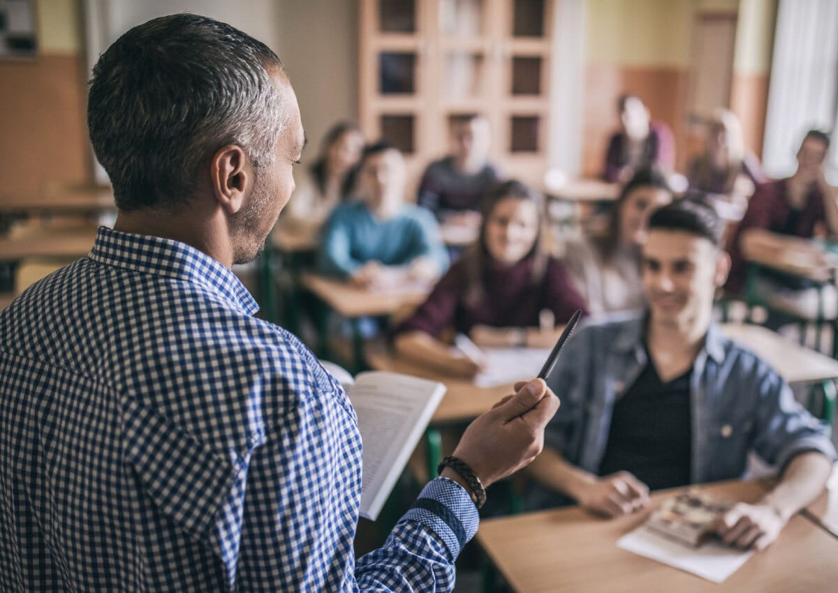 A male teacher stands in front of a classroom of older students.