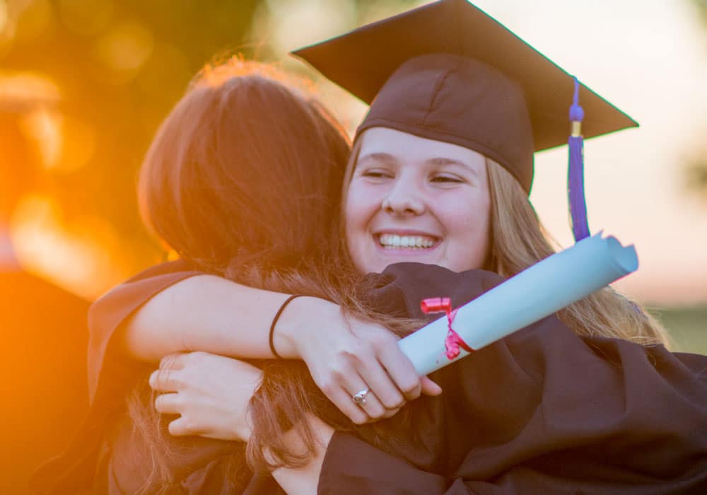 A girl in a graduation cap and holding a diploma hugs another girl.
