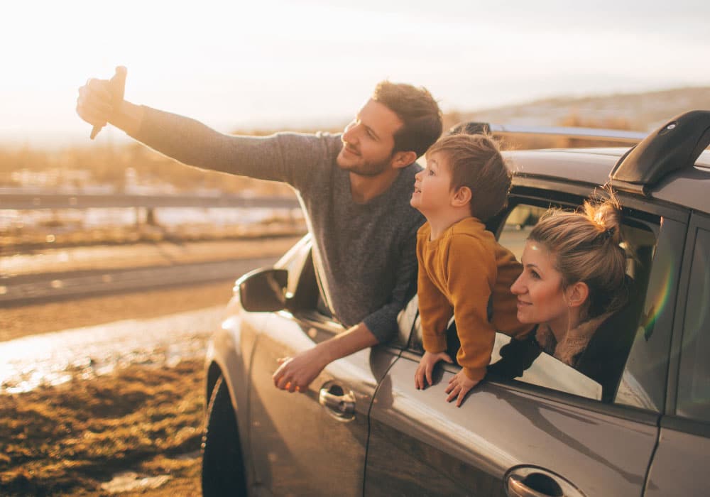 A family taking a selfie while in a car, they are hanging out the window of a parked car.