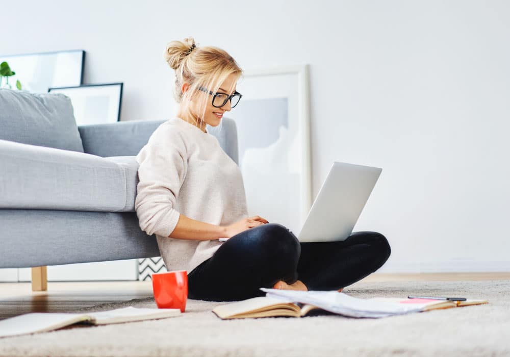 Female sits on floor on laptop with notebooks open next to her