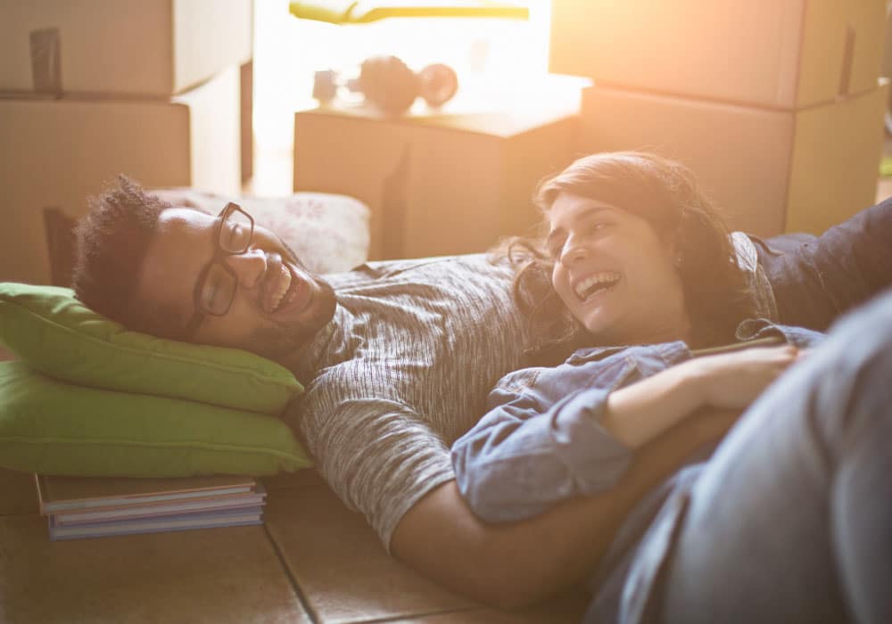 a couple lays on a floor and is laughing, there are moving boxes behind them.