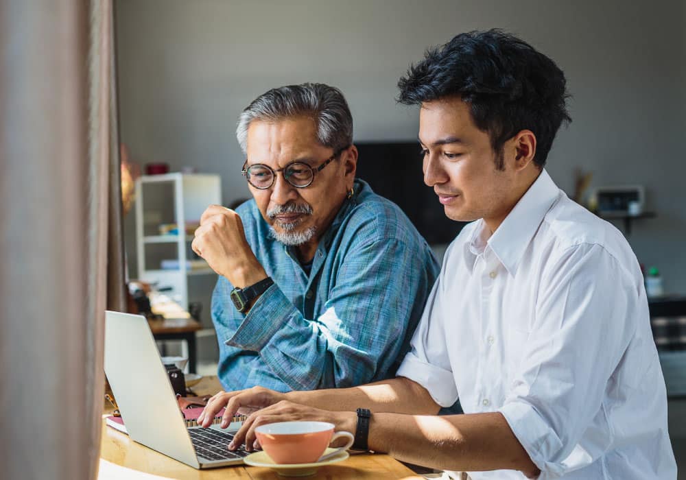 Two people sit at a desk looking at a laptop while one types on it.