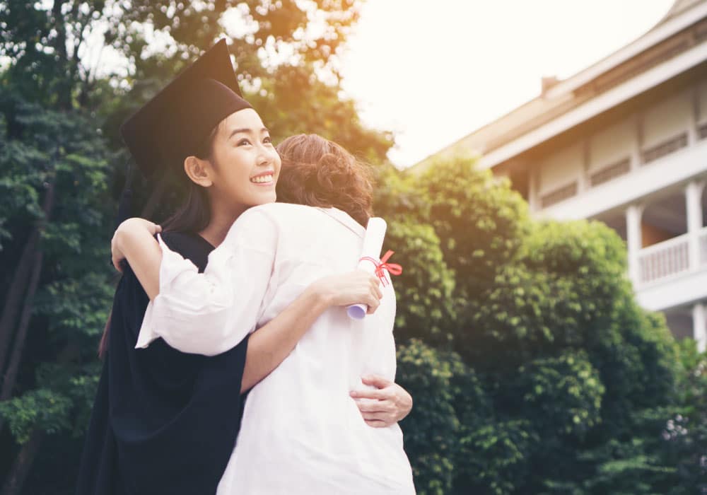 A woman with a graduation cap and gown smiles and hugs another woman.