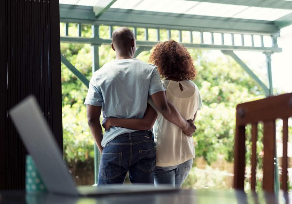 A couple standing on a porch, their backs are to us, and there is an open laptop on the table in front of them.