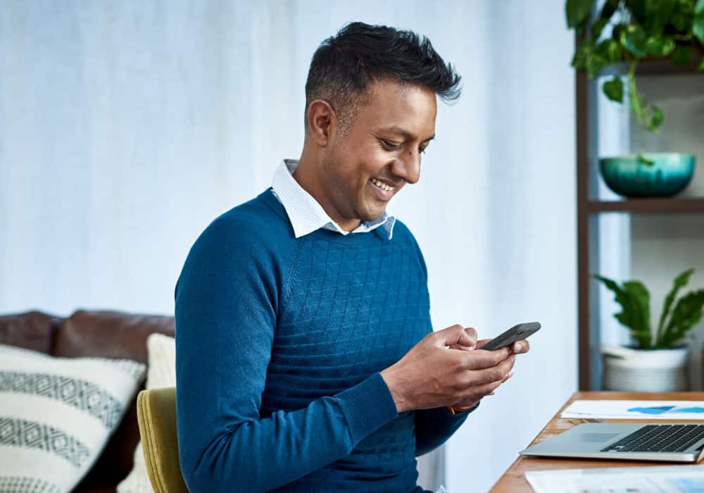 A man sits in a living room and looks down at a phone and smiles.