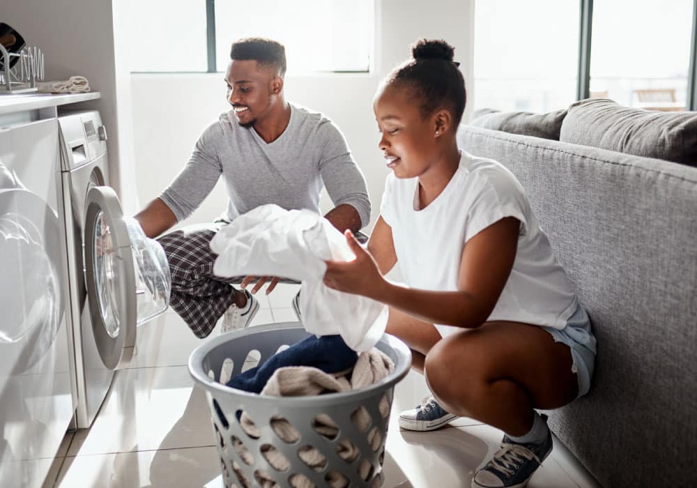 A couple doing laundry in a home. The man using the washing machine and the woman is folding laundry.