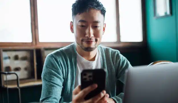 Man sits at table holding his mobile phone and also has his laptop open.