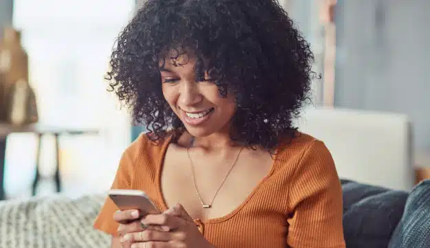A woman sitting on a couch and looking down at a cell phone, she is smiling.