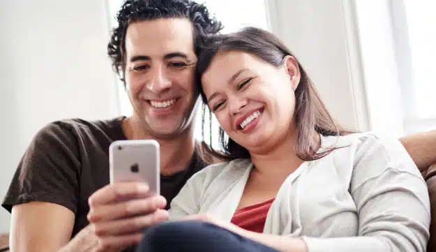 A couple sits on a couch and looks at the screen of a cell phone together. They are laughing.