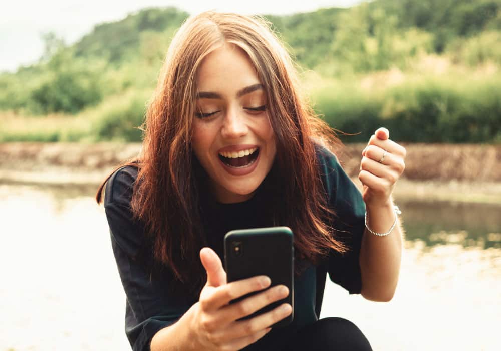 A woman standing on the edge of a river or lake and using a cell phone. She is smiling.