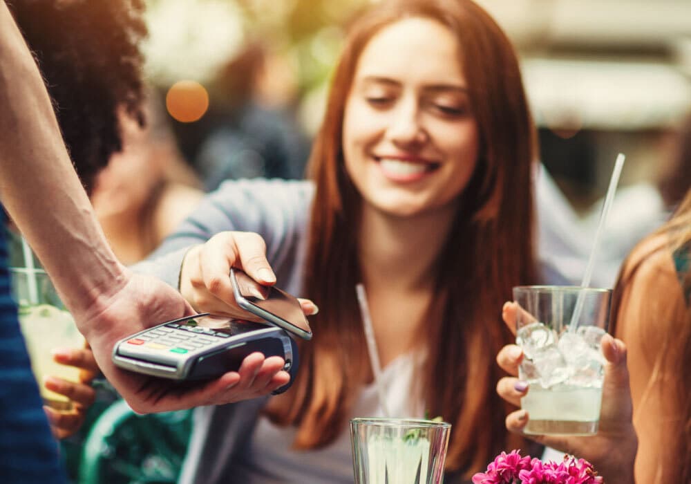 A woman using a cell phone to pay, she has drinks in front of her.