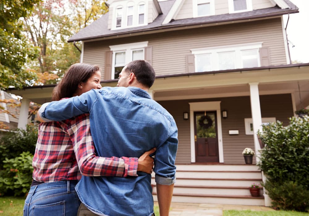 A couple standing outside of a house. they are looking at each other and smiling.