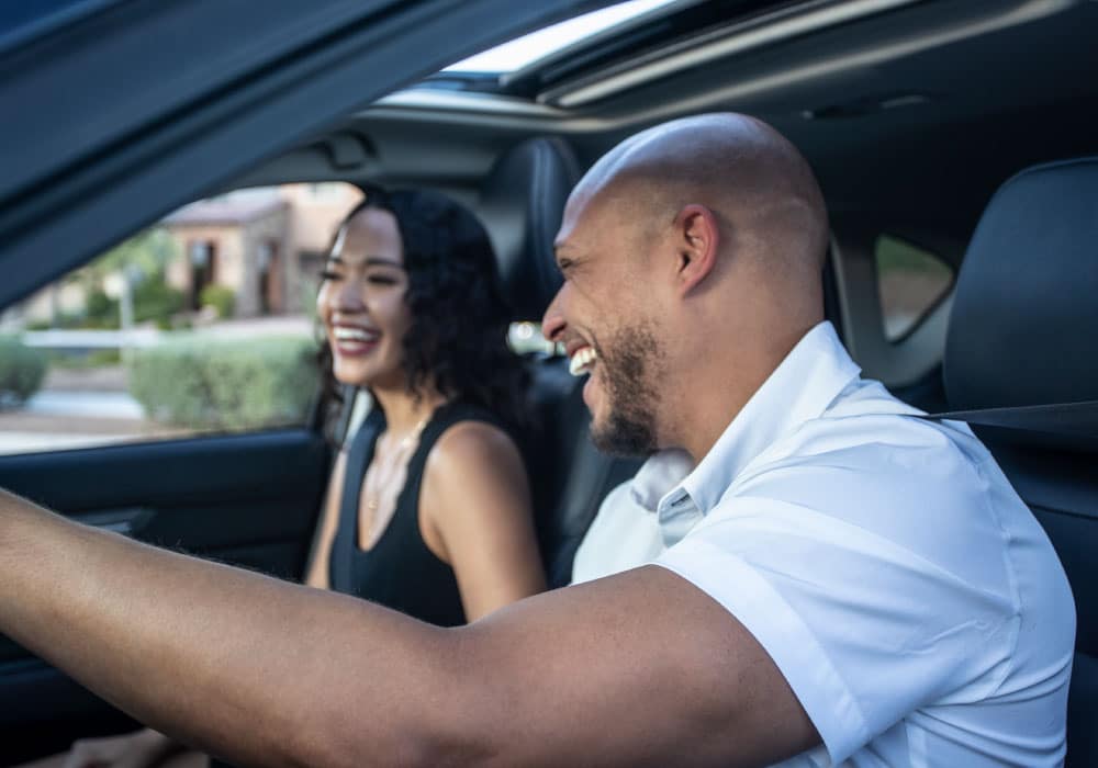 A man and a woman in a car, the man is smiling and driving and looking over at the woman.