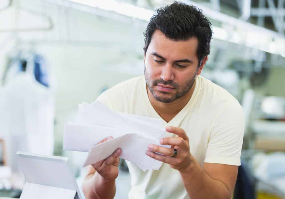 A man leaning over a table and looking through some bills. He has a tablet on a table in front of him.