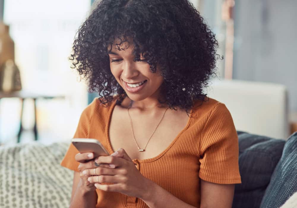 A woman sitting on a couch and smiling down at a cell phone.