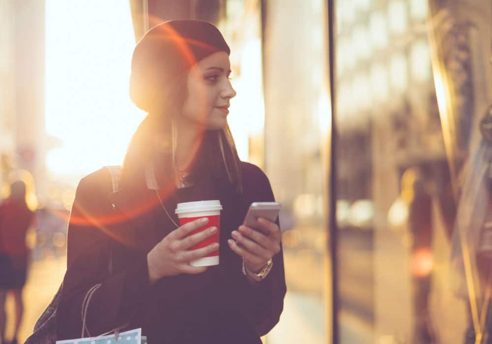 A woman walks outside a window with a cup of coffee and a cell phone.