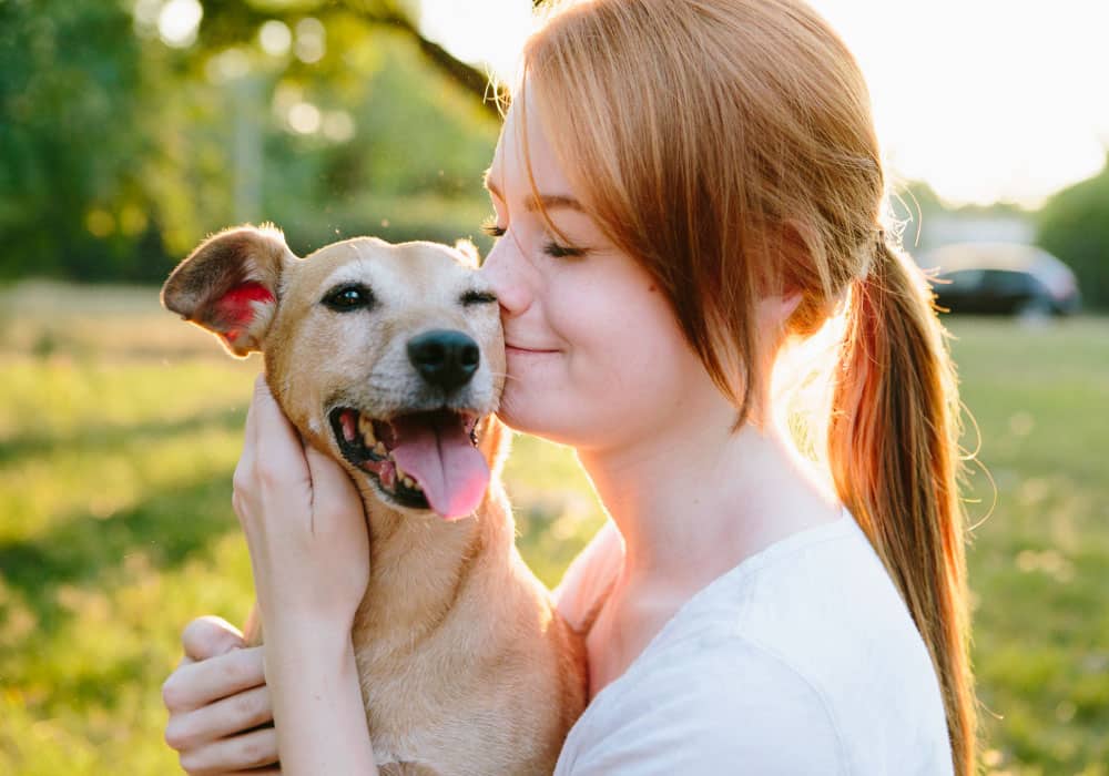 Woman hugs and kisses dog’s face while outside.