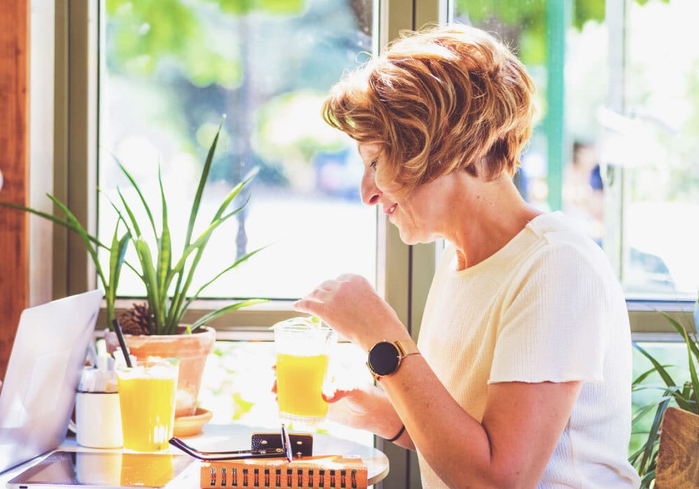 woman, holding a glass of orange juice, sits at a table in front of windows