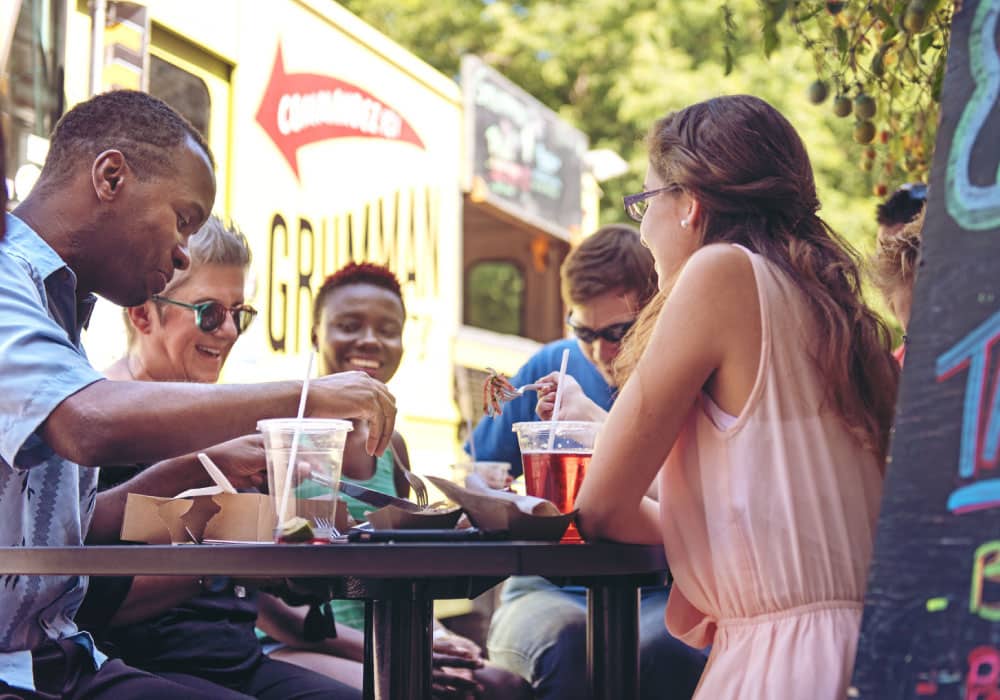Group of people sit outside in front of food truck laughing and eating.