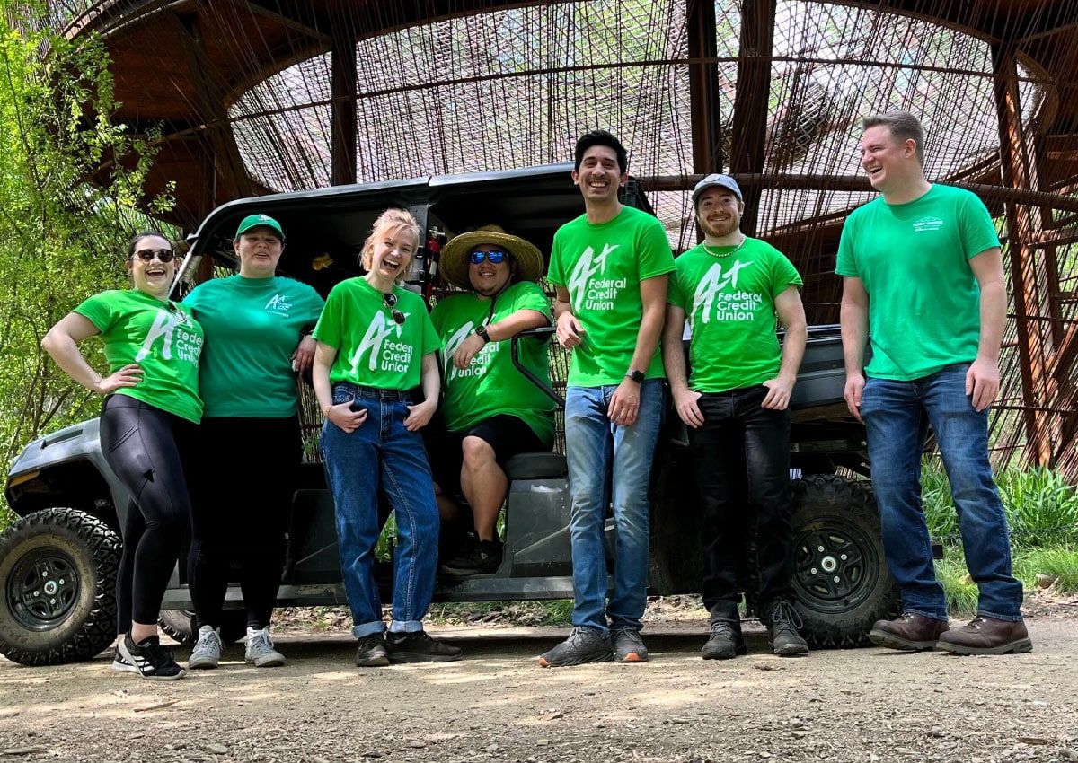 A+FCU Team members wearing green shirts, standing outside in front of a side-by-side.