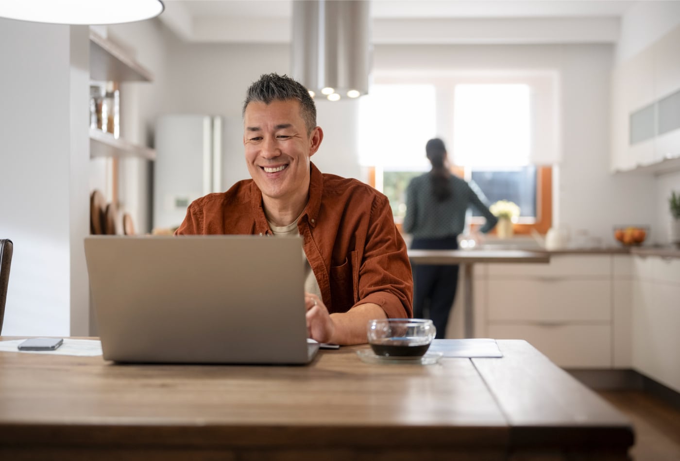 Man sits at kitchen table working on laptop and smiling.