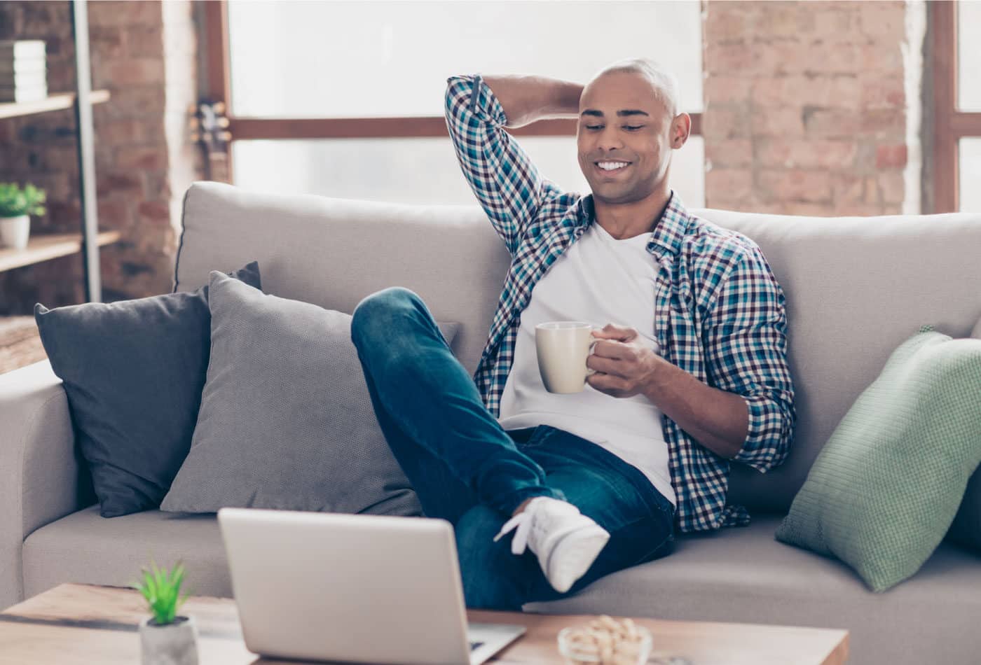 Man sitting on a couch in front of a laptop with his legs crossed while holding a coffee mug