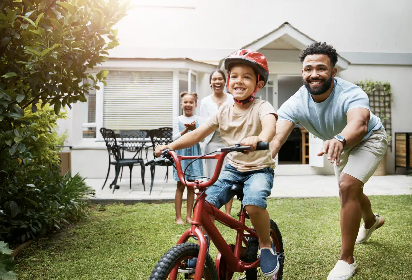 Family of four in the backyard while the dad helps the younger son ride a bike. Mom and sister clap in the back.