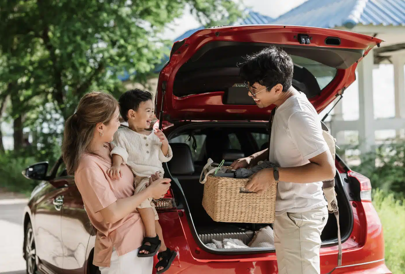 Mother holding her toddler while a man loads a woven basket from the back of a red SUV with the trunk open on a sunny day, surrounded by greenery.