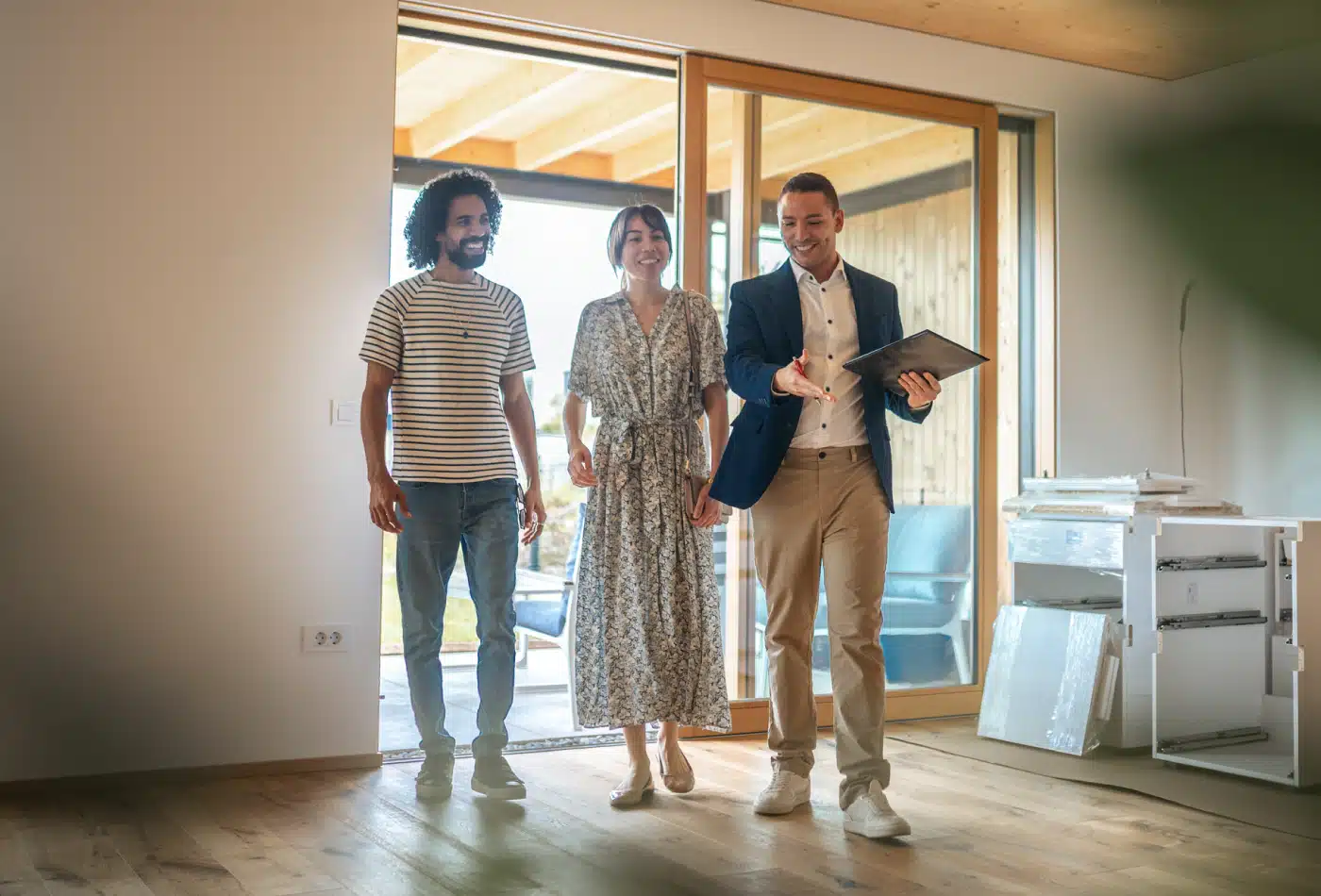 Real estate agent in a blazer guides a smiling couple through a sunlit living room with large glass doors.