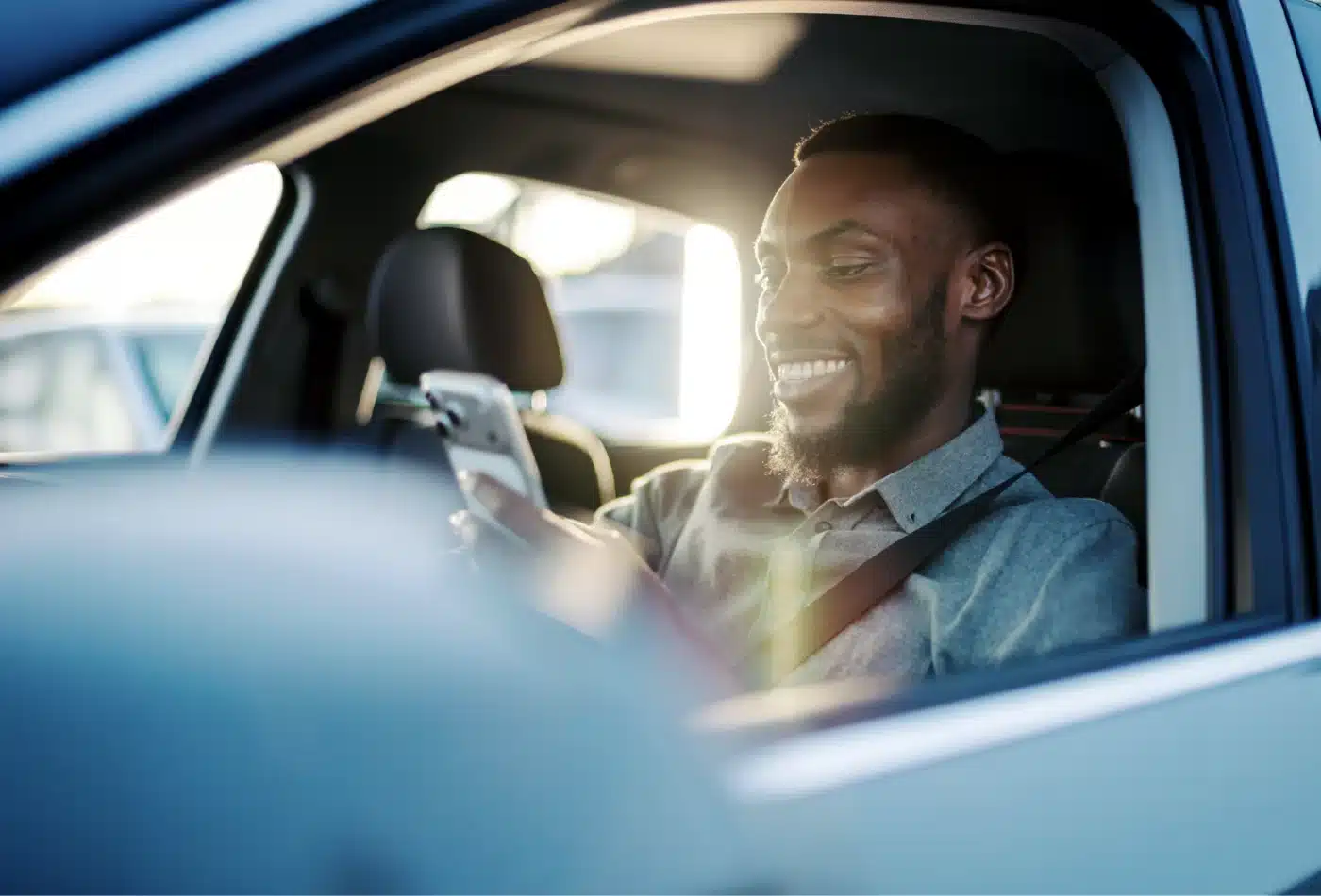 A man wearing a seat belt smiles while looking at his cell phone from the driver's seat of a car.