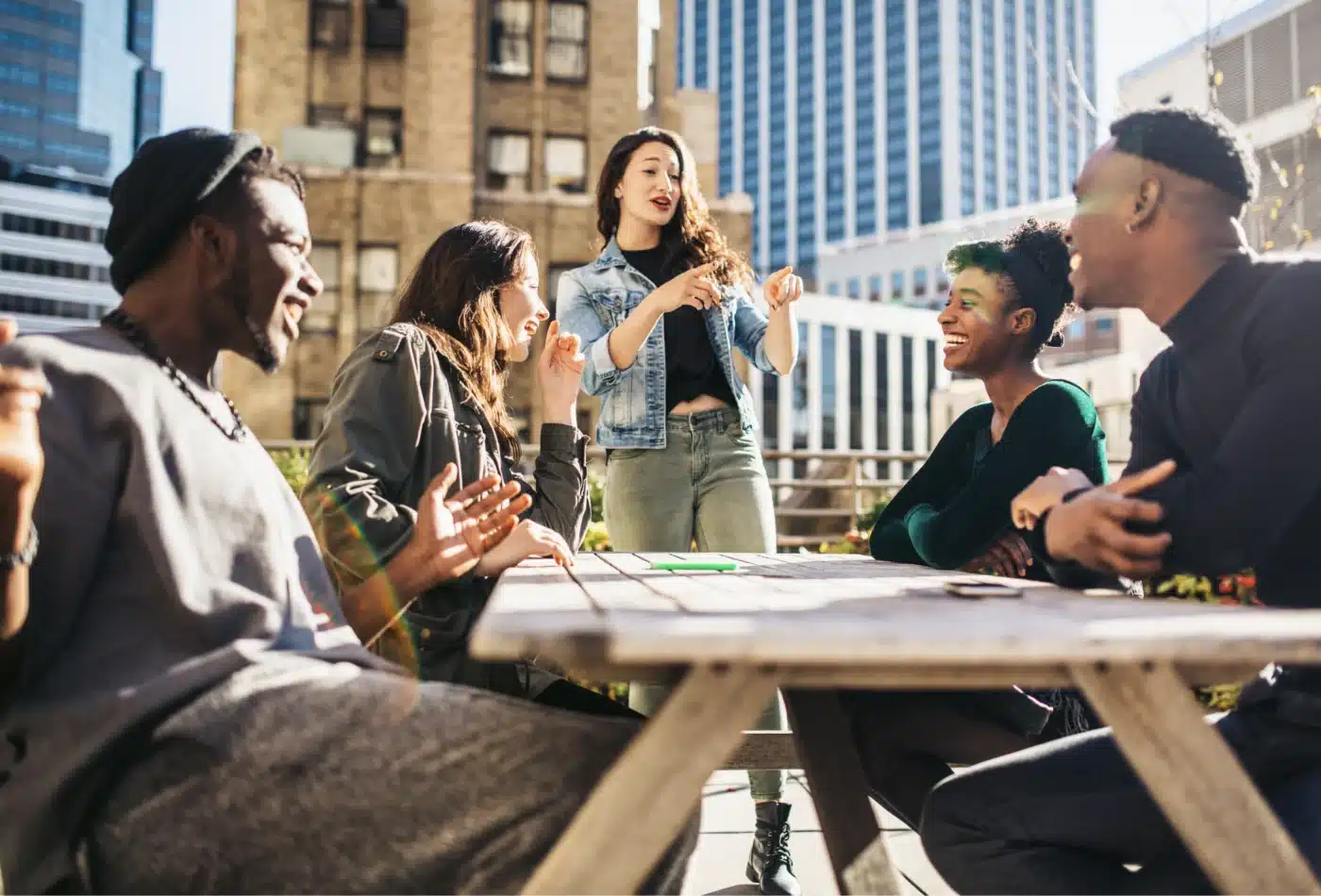 A group of diverse friends sitting around a wooden outdoor table in an urban setting, chatting and laughing as one person speaks standing up.
