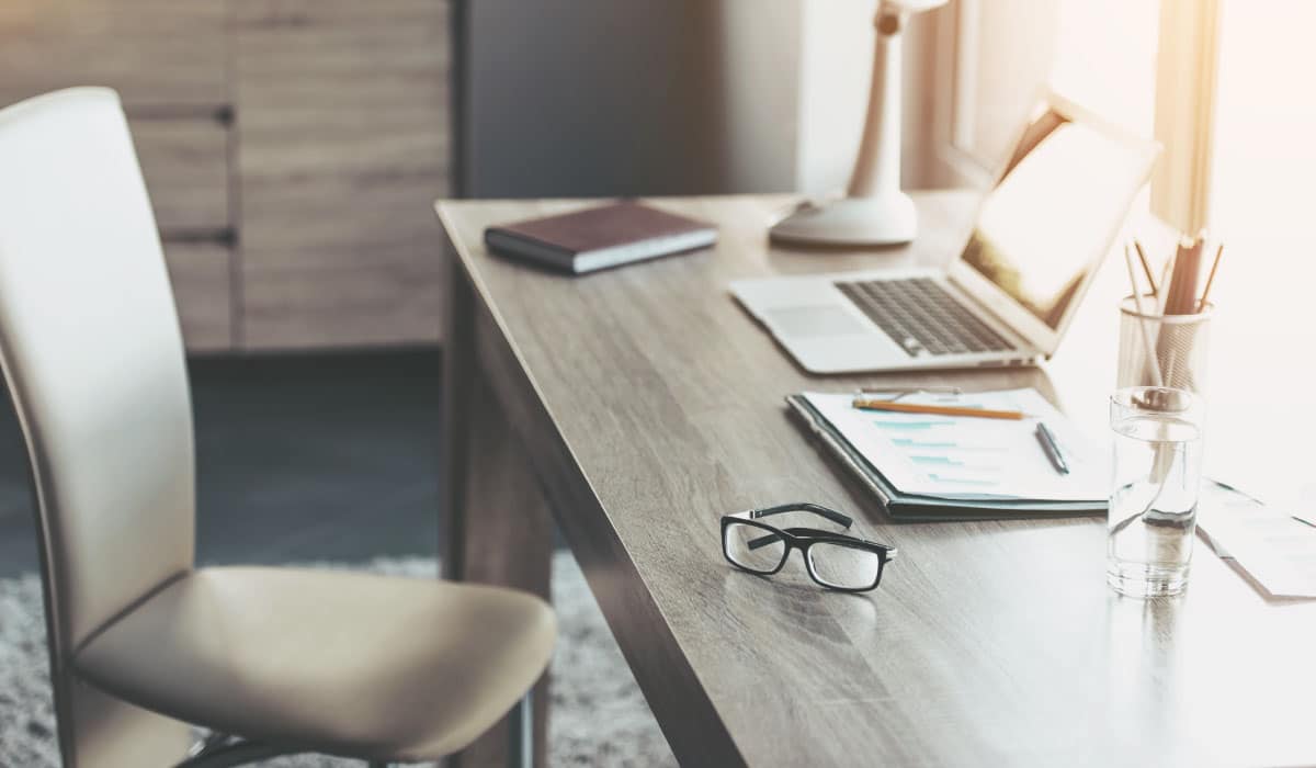 A work desk with a laptop, notebook, and reading glasses on top. An empty chair is pushed away from the desk.