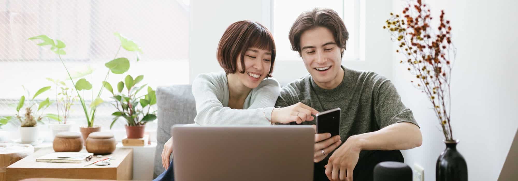 A couple sits together in a house, the man is holding a cell phone and the woman is pointing to something on the screen.