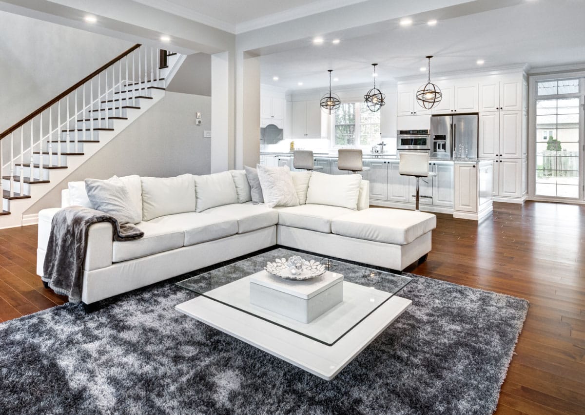 Staged living room and kitchen with all white furniture and a gray rug, staircase off to the side.