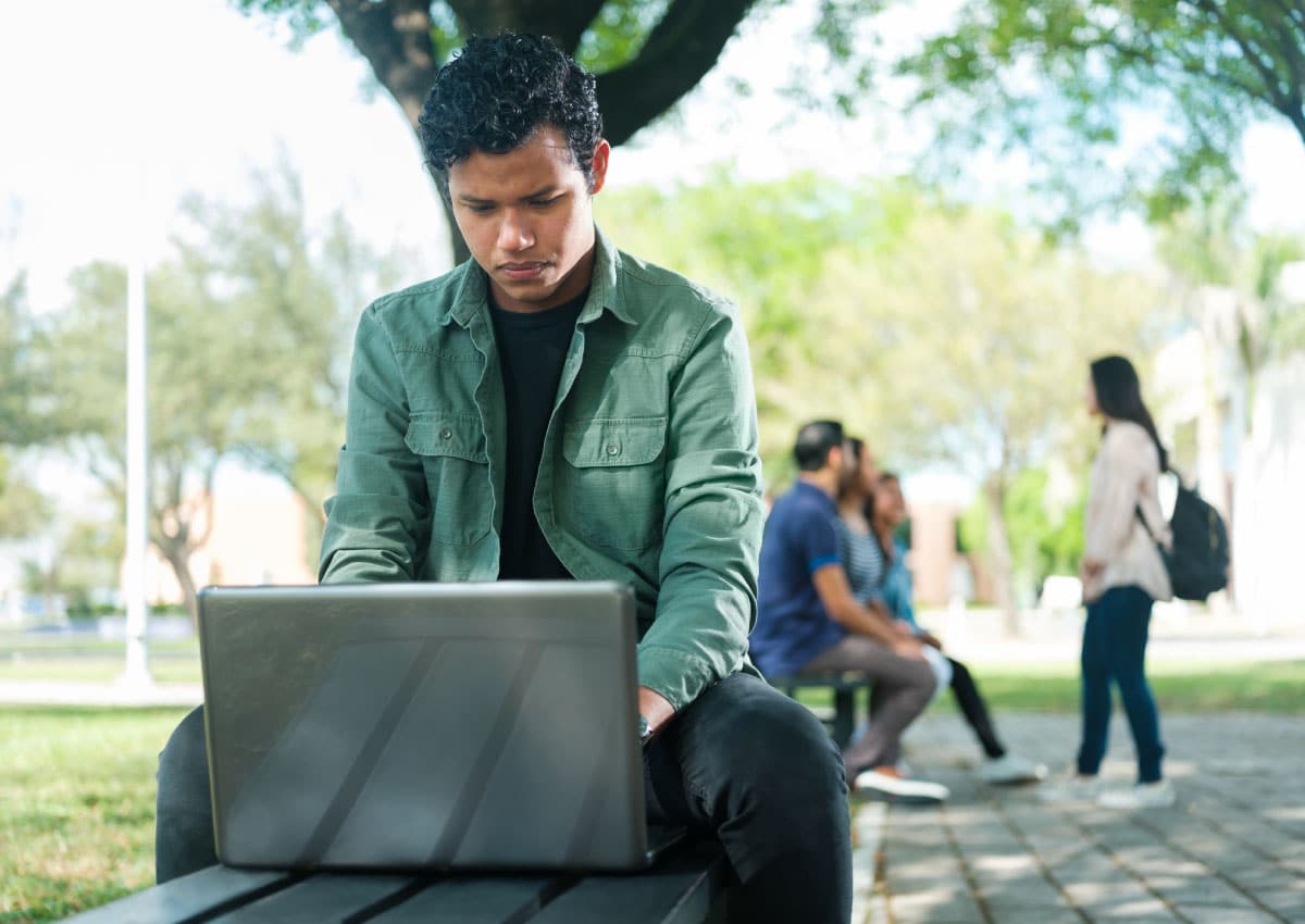 Young man sits outside under a tree working on a laptop. A group of people are talking in the background.