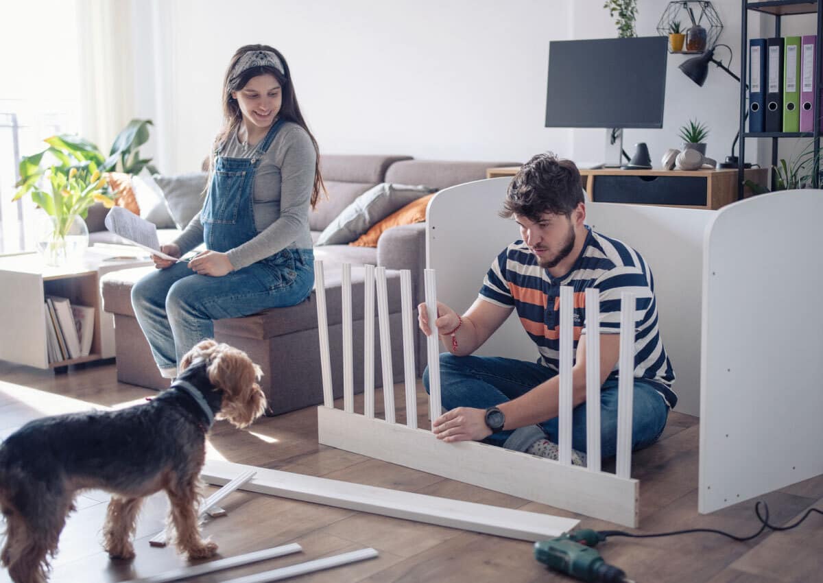 Pregnant woman sits on couch holding directions and man sits on floor putting together a crib while dog watches