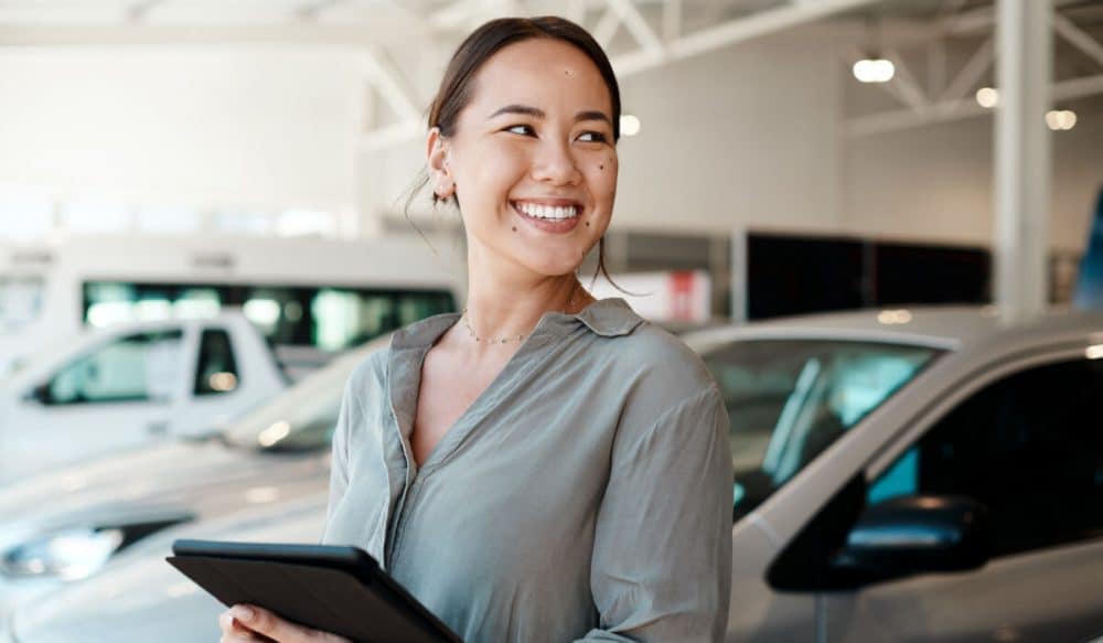 A woman stands in front of several cars, holding a tablet.