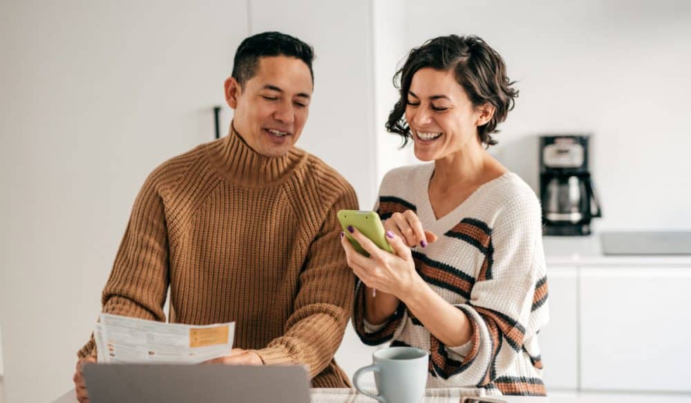 a couple standing at a kitchen island. The woman is showing the man something on a cell phone, there is a laptop open in front of them.