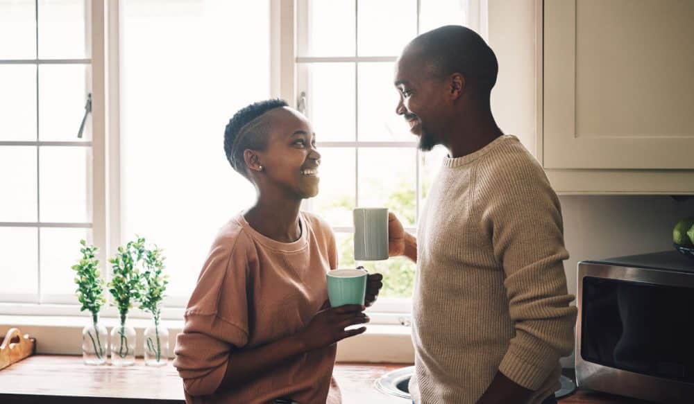 A couple standing in a kitchen, they are laughing and holding mugs.