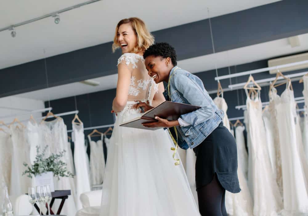 A woman tries on a wedding dress with another woman taking her measurements, they are laughing