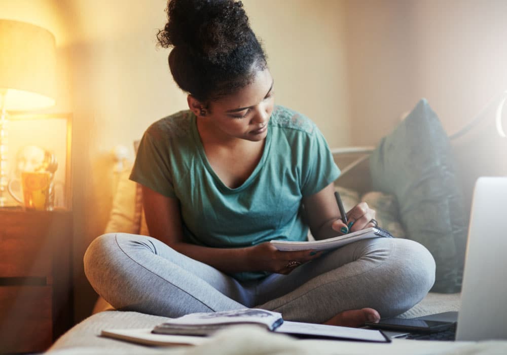 Young adult female sits cross-legged on couch while writing in a notebook