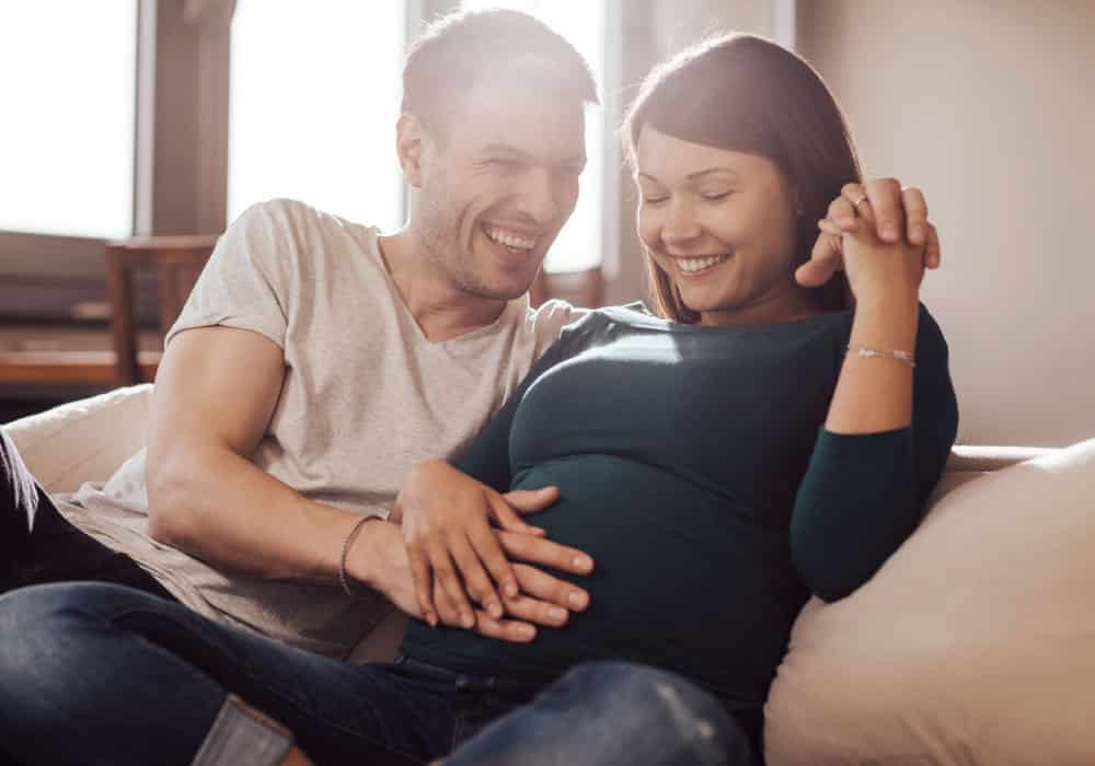A couple sits on a couch and laughs, the woman is pregnant