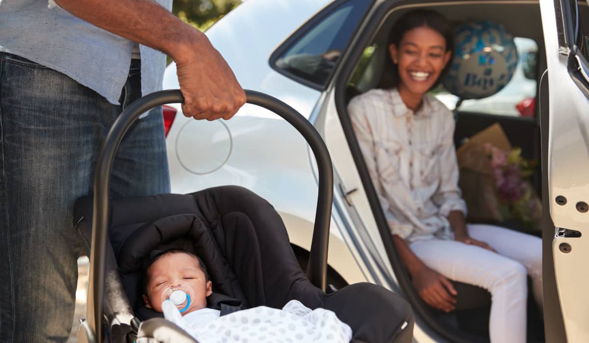 A man holds a newborn baby in a car seat while a woman sits in the car and smiles