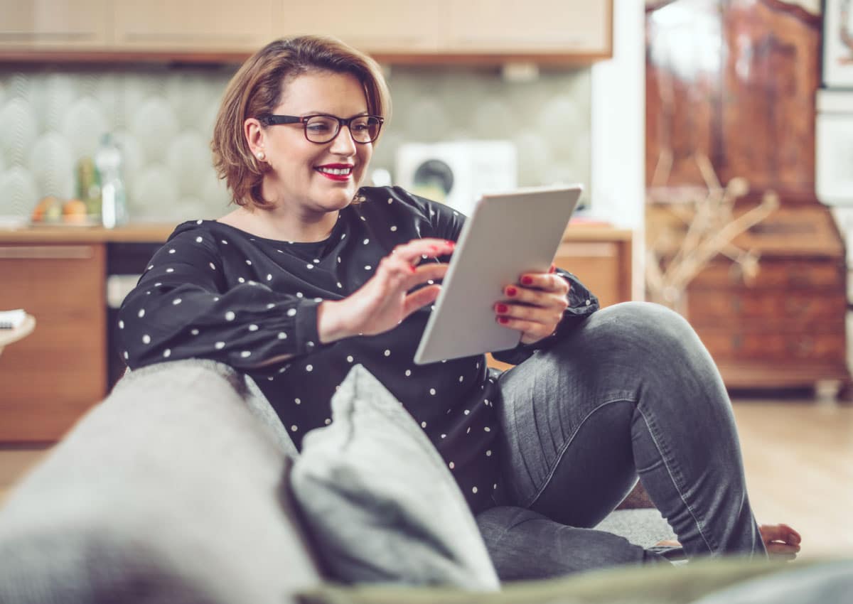 Woman sits on couch looking at tablet while smiling